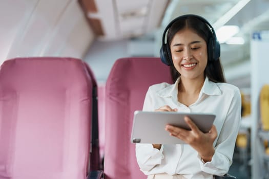 Young Asian attractive business woman passenger sitting on business class luxury plane while wearing wireless headphone for relax and working with tablet during the flight.