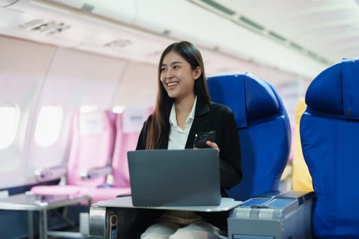 Asian business woman passenger sitting on business class luxury plane while working using smart phone mobile talking and use laptop computer with simulated space using on board wireless connection