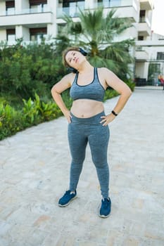 Adorable fat woman in tracksuit is engaged in fitness outdoor side view portrait. Young overweight woman lunges outdoors on warm summer day. Healthy lifestyle and weight loss.