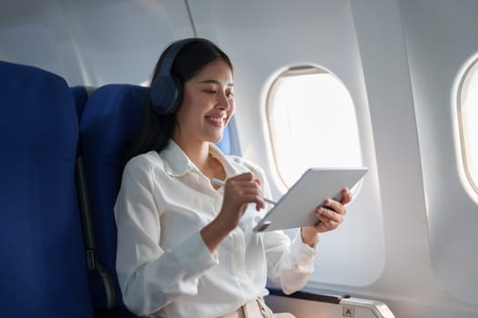 Young Asian attractive business woman passenger sitting on business class luxury plane while wearing wireless headphone for relax and working with tablet during the flight.