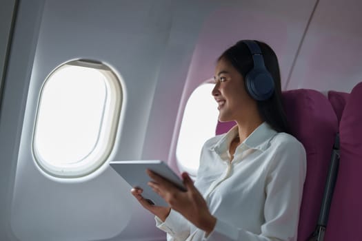 Young Asian attractive business woman passenger sitting on business class luxury plane while wearing wireless headphone for relax and working with tablet during the flight.