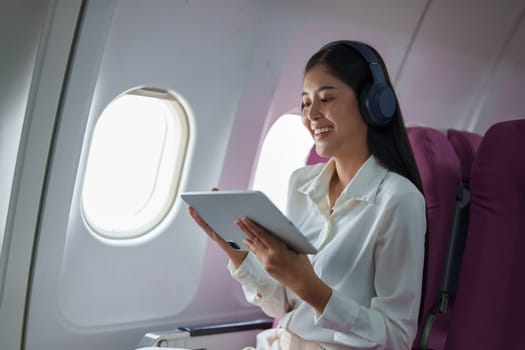 Young Asian attractive business woman passenger sitting on business class luxury plane while wearing wireless headphone for relax and working with tablet during the flight.