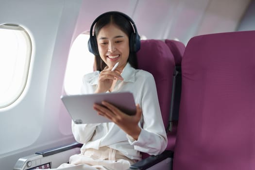 Young Asian attractive business woman passenger sitting on business class luxury plane while wearing wireless headphone for relax and working with tablet during the flight.