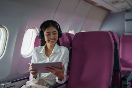 Young Asian attractive business woman passenger sitting on business class luxury plane while wearing wireless headphone for relax and working with tablet during the flight.