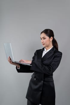 Confident young businesswoman stands on isolated background, working on laptop posing in formal black suit. Office lady or manager with smart and professional appearance. Enthusiastic