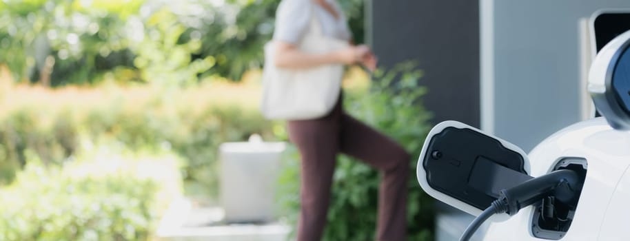 Focus EV charger plugged into EV car at home charging station with blurred background of progressive woman walking in background. Elective vehicle powered by clean energy for eco-friendly concept.