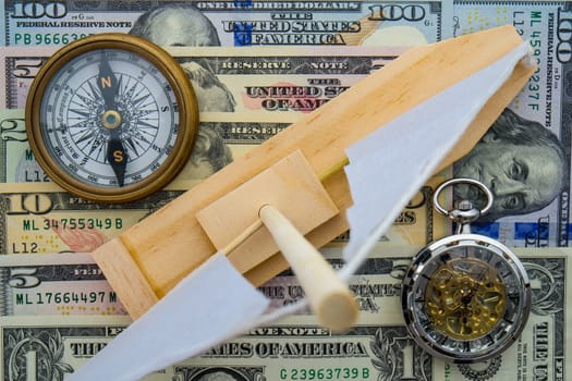 Toy sailboat, compass, and pocket watch sitting on US currency bills of different denominations showing concept high cost of leisure boating, time, direction