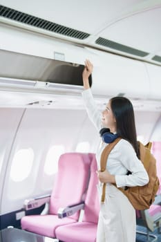 Young asian attractive woman travel by airplane, Passenger wearing headphone putting hand baggage in lockers above seats of plane.
