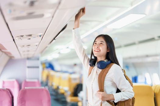 Young asian attractive woman travel by airplane, Passenger wearing headphone putting hand baggage in lockers above seats of plane.