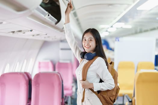 Young asian attractive woman travel by airplane, Passenger wearing headphone putting hand baggage in lockers above seats of plane.