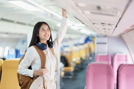 Young asian attractive woman travel by airplane, Passenger wearing headphone putting hand baggage in lockers above seats of plane.