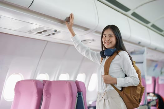 Young asian attractive woman travel by airplane, Passenger wearing headphone putting hand baggage in lockers above seats of plane.