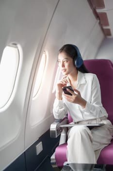 Young Asian attractive business woman passenger sitting on business class luxury plane while wearing wireless headphone and using smart phone mobile for relax during the flight.