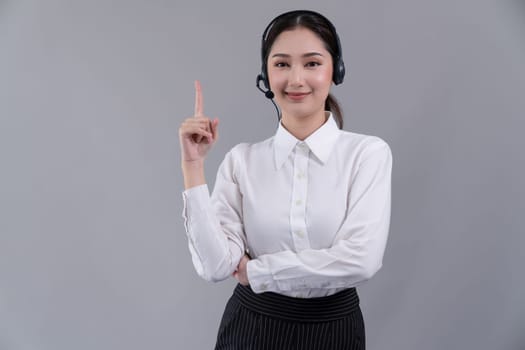 Asian female call center operator with smile face advertises job opportunity, wearing a formal suit and headset pointing finger for product on customizable isolated background. Enthusiastic