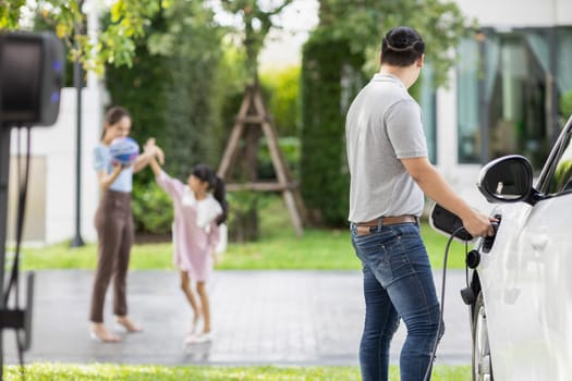 Focus image of progressive man charging electric car from home charging station with blur mother and daughter playing together in the background.