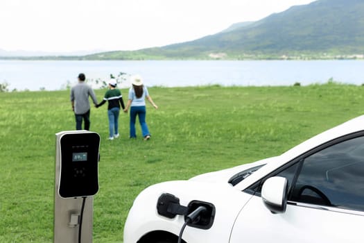 Concept of progressive happy family enjoying their time at green field and lake with electric vehicle. Electric vehicle driven by clean renewable from eco-friendly power sauce.