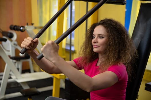 Lifting weights at a gym. A well-equipped exercise room. Workout performed at the gym. A woman uses sports equipment to exercise her upper body. Performing weight training exercises.