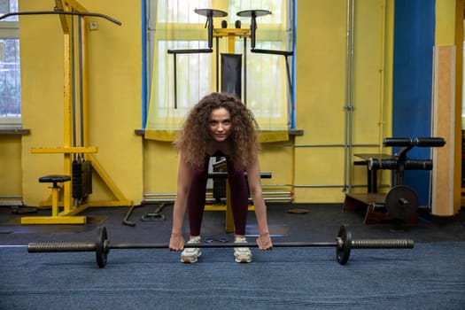 A girl with curly hair exercises at the gym. Lifting weights attached to a barbell. Exercising arm and back muscles with equipment available at the gym. An athletic girl keeps in shape by exercising.