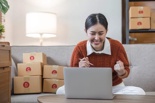 Asian business woman leaning on the sofa use laptop computer checking customer order online shipping boxes at home. Starting Small business entrepreneur SME. Online business, Work at home concept...