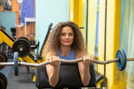 Front view of a girl lying on her stomach on an exercise bench. The woman is holding an exercise barbell and lifting it while exercising her biceps. Blurred background. Other exercise equipment can be seen in the background.