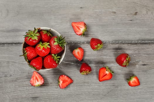 Tabletop view - small ceramic bowl of strawberries, more cut fruits lying around on gray wood desk.
