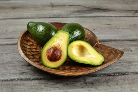 Avocados in wooden carved bowl, one of them cut in half, seed visible.