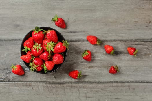 Tabletop view, small black ceramic bowl with strawberries, some of them spilled next to it on gray wood desk.