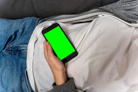 an unrecognizable girl uses a smartphone with a green screen while lying at home on the couch. a smartphone with a chromakey in female hands. View videos and photos on social networks. web browsing on the Internet, browsing. Dependence on gadgets