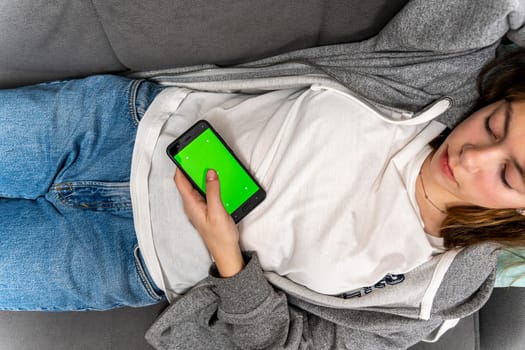 an unrecognizable girl uses a smartphone with a green screen while lying at home on the couch. a smartphone with a chromakey in female hands. View videos and photos on social networks. web browsing on the Internet, browsing. Dependence on gadgets