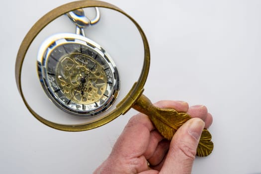 A man's hand holding a gold handled magnifying glass examining a pocket watch with visible gears. Isolated on white background. Concept time management. High quality photo