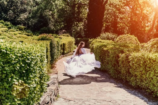 Brunette runs white dress park. A beautiful woman with long brown hair and a long white dress runs along the path along the beautiful bushes in the park, rear view.