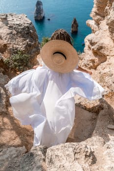 Woman white dress sea rocks. A girl in a hat and a long white dress descends the stairs between the yellow rocks overlooking the sea. The stone can be seen in the sea. Travel photograph