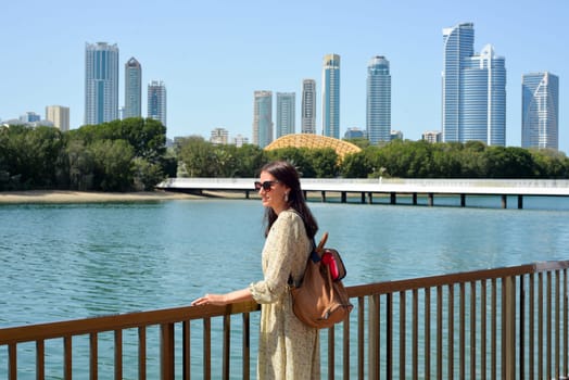 The skyline of Sharjah is visible from the waterfront - a woman tourist with a backpack enjoys the view of the water and skyscrapers. Cityscape of Sharjah UAE