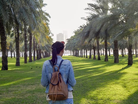 A woman traveler in a denim jacket with a backpack in a palm garden in Sharjah, UAE. Which is located on the Sharjah Corniche