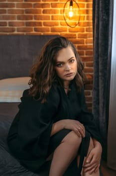 Portrait of a beautiful woman in a black bathrobe, who is sitting on the bed, looking at the camera.