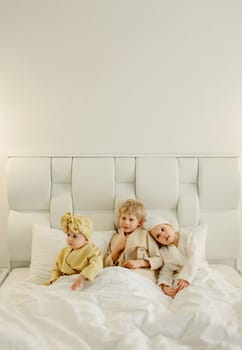 Children lie in a white bed in bathrobes after taking a bath, smiling and looking at the camera. View from above.