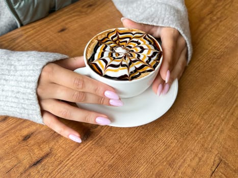 Girl 's hands are holding white cup of coffee with spider web pattern on top of foam, latte art, against background of wooden table, close-up