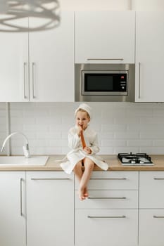 A girl in a white coat sits on the kitchen table and eats marmalade.