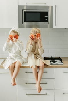 A girl and a boy in bathrobes sit in the kitchen and close their eyes with candied oranges.