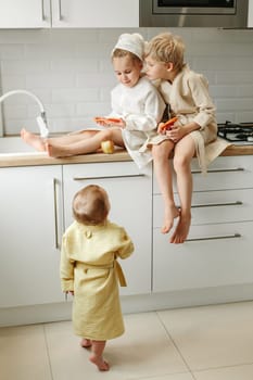 A girl and a boy in bathrobes are sitting in the kitchen and holding candied oranges in their hands, the youngest child is looking at them.