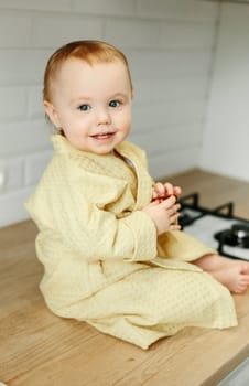 A girl in a yellow bathrobe sits on the kitchen table and eats marmalade.