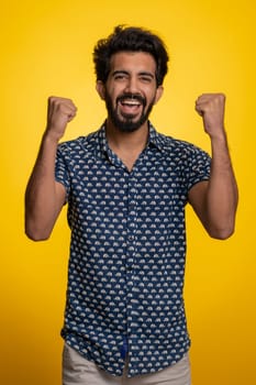 Happy joyful indian man shouting, raising fists in gesture I did it, celebrating success, winning, birthday, lottery jackpot goal achievemen, good news. Young hindu guy on yellow studio background