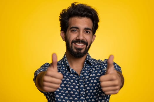 Like. Indian young man in shirt raises thumbs up agrees with something, gives positive reply recommends advertisement likes good. Handsome bearded hindu guy isolated alone on yellow studio background