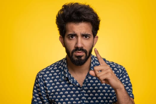 Quarrel. Displeased young indian man gesturing hands with irritation and displeasure, blaming scolding for failure, asking why this happened. Bearded hindu guy isolated on yellow studio background