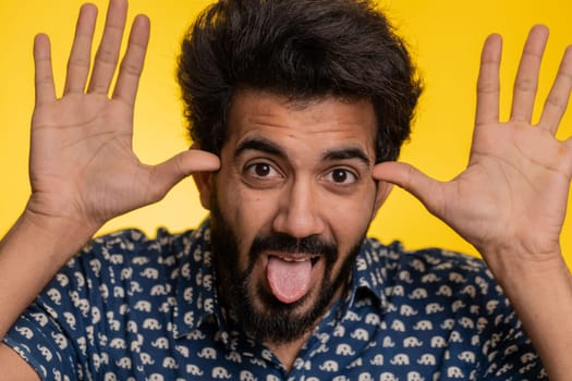 Young indian man showing tongue making childish faces at camera, fooling around, joking, aping with silly face, teasing, bullying abuse. Handsome hindu guy isolated on studio yellow background indoors