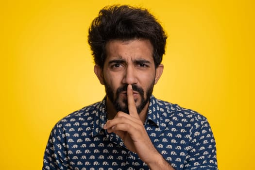 Shh be quiet please. Young indian man presses index finger to lips makes silence gesture sign do not tells secret, stop gossip. Handsome bearded hindu guy isolated alone on yellow studio background