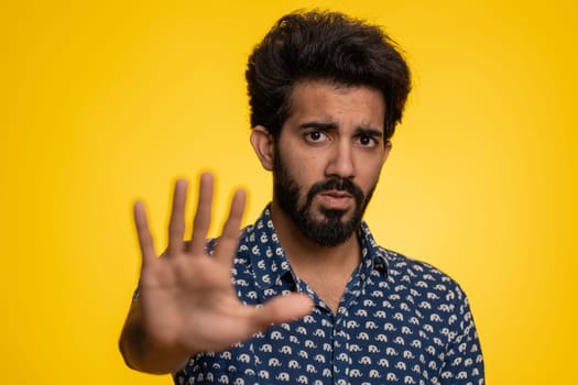 Indian man say No hold palm folded crossed hands in stop gesture, warning of finish, prohibited access, declining communication, body language, danger. Young guy on yellow background. People lifestyle