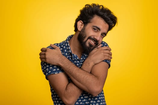 Come to me, I want to embrace you. Indian handsome man spread hands and give hug to you. Pleasant expression, love feelings. Young guy isolated alone on yellow studio background. People lifestyles