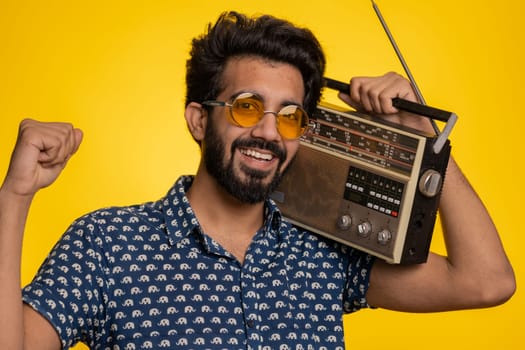 Indian man using retro tape record player to listen music, disco dancing of favorite track, having fun, entertaining, fan of vintage technologies. Handsome hindu guy isolated on yellow background