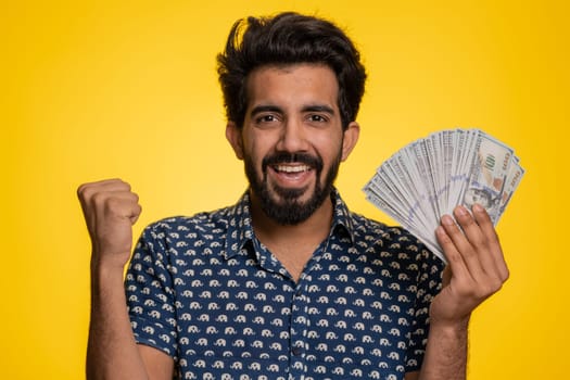 Rich pleased boss hindu man waving money dollar cash banknotes bills like a fan, success business career, lottery winner, big income, wealth. Handsome indian guy isolated on yellow background, indoor